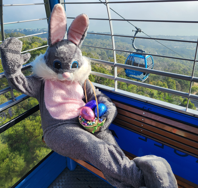 Easter bunny sits in a blue gondola holding a basket of easter eggs. Another gondola flies past